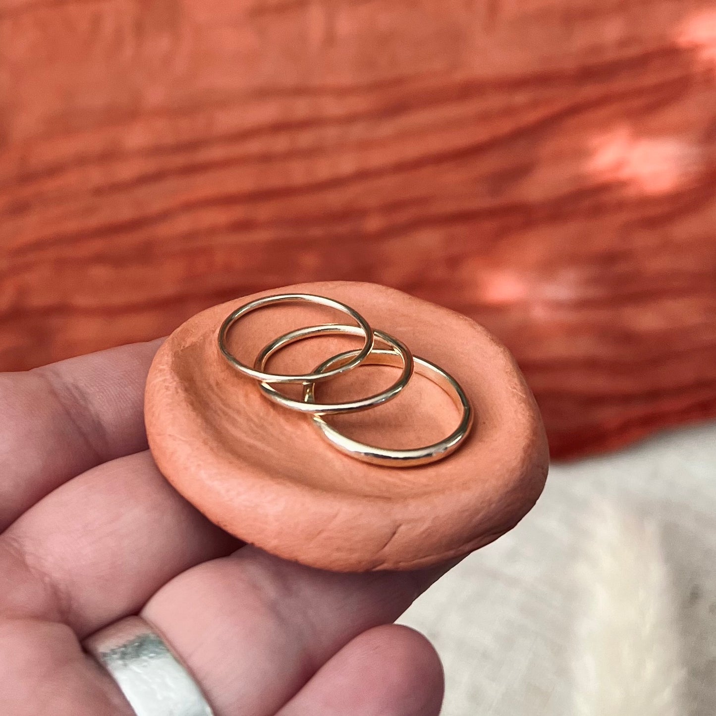A stack of three recycled gold rings in different textures and sizes, held in a person's hand, with a bohemian-style orange fabric in the background.