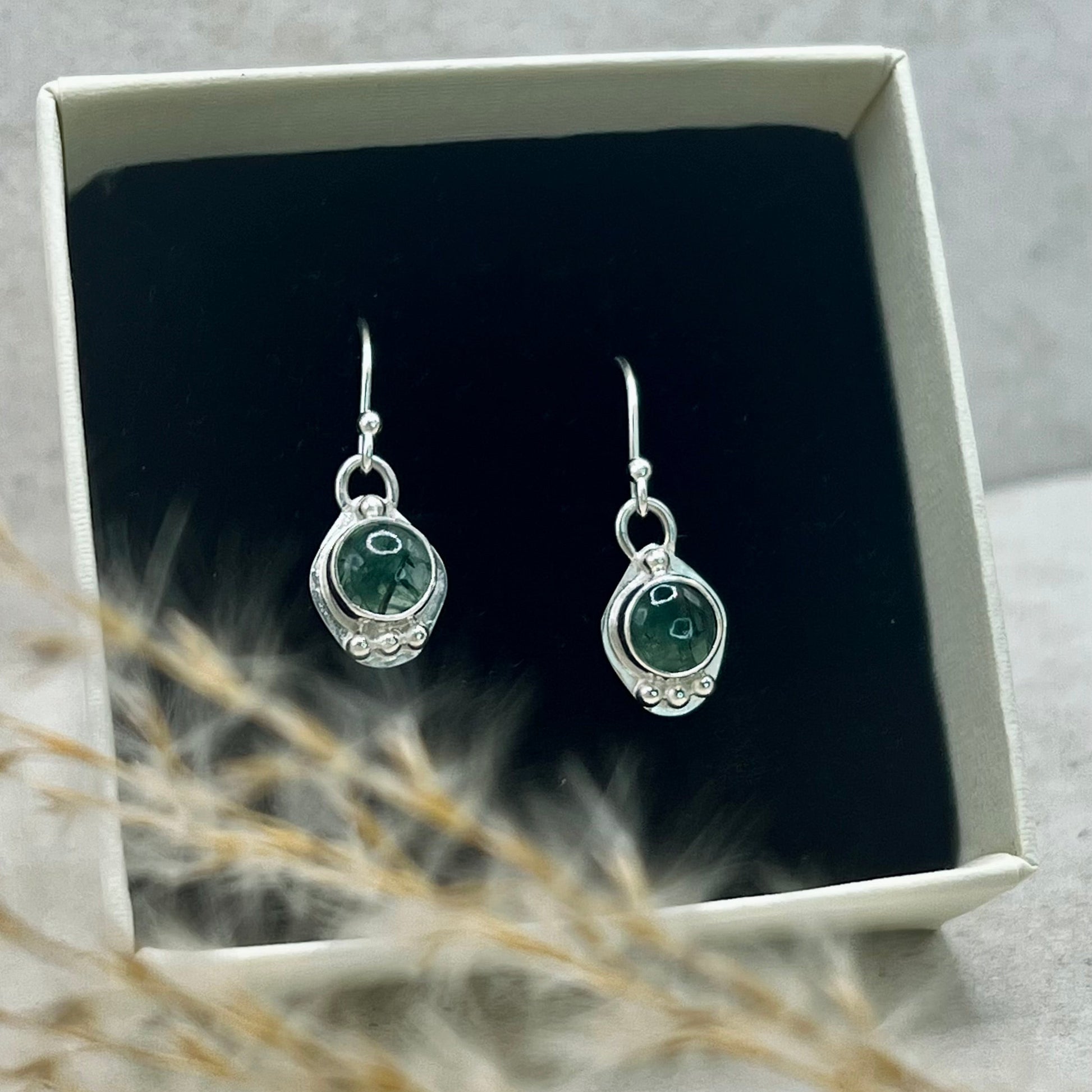 Silver earrings with green stones in a white box on a gray background