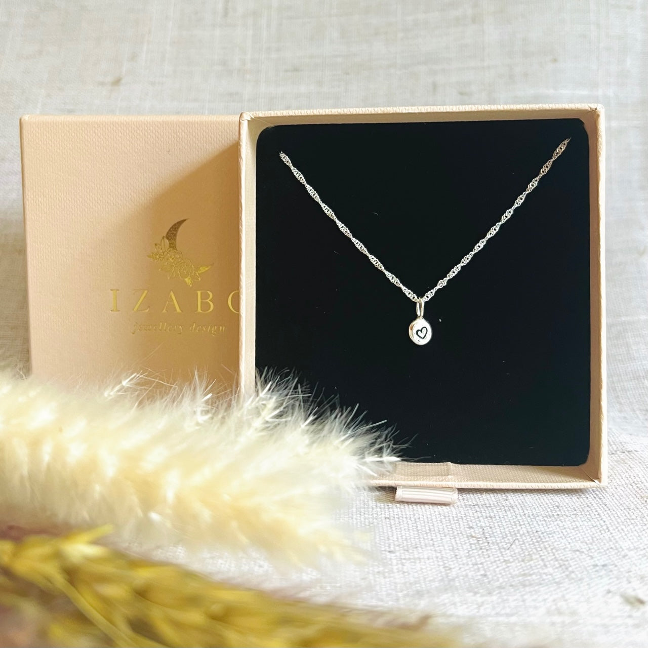 Beautiful little recycled silver nugget necklace on a twisted rope chain, placed in a cream coloured IZABO branded match box style jewellery gift box. There are some dry foliage in front of the box, on a sand coloured linen type table cover.