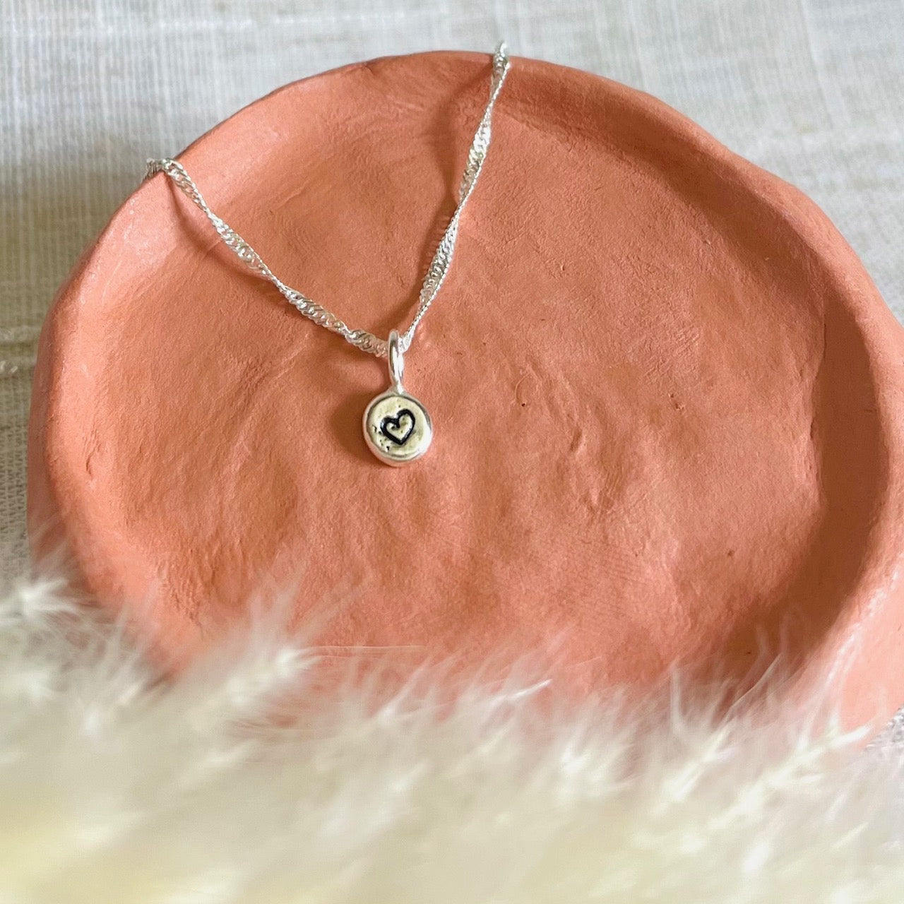 Cute little recycled silver nugget with a hearth motif, on a twisted silver chain. Placed on a terracotta dish with pampas grass in front, on a sand coloured linen type table cover.