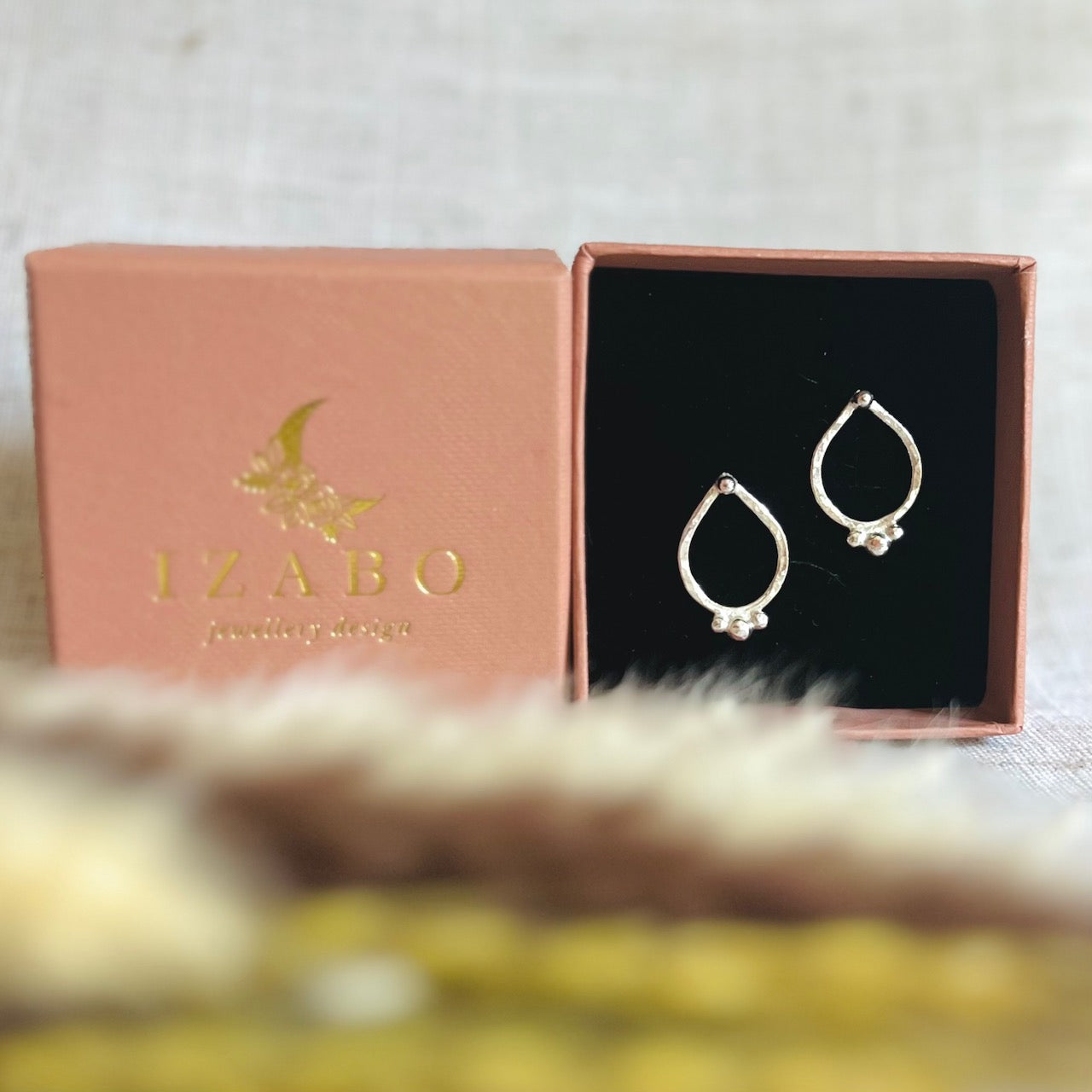 recycled silver lotus or tear drop earrings with little silver balls, one on the top and 3 little ones at the bottom or the drop in a line. the earrings are in their terracotta coloured gift box which has a metallic gold IZABO writing with a moon and flower logo. the gift box is placed on a cream table cover behind some dry foliage.