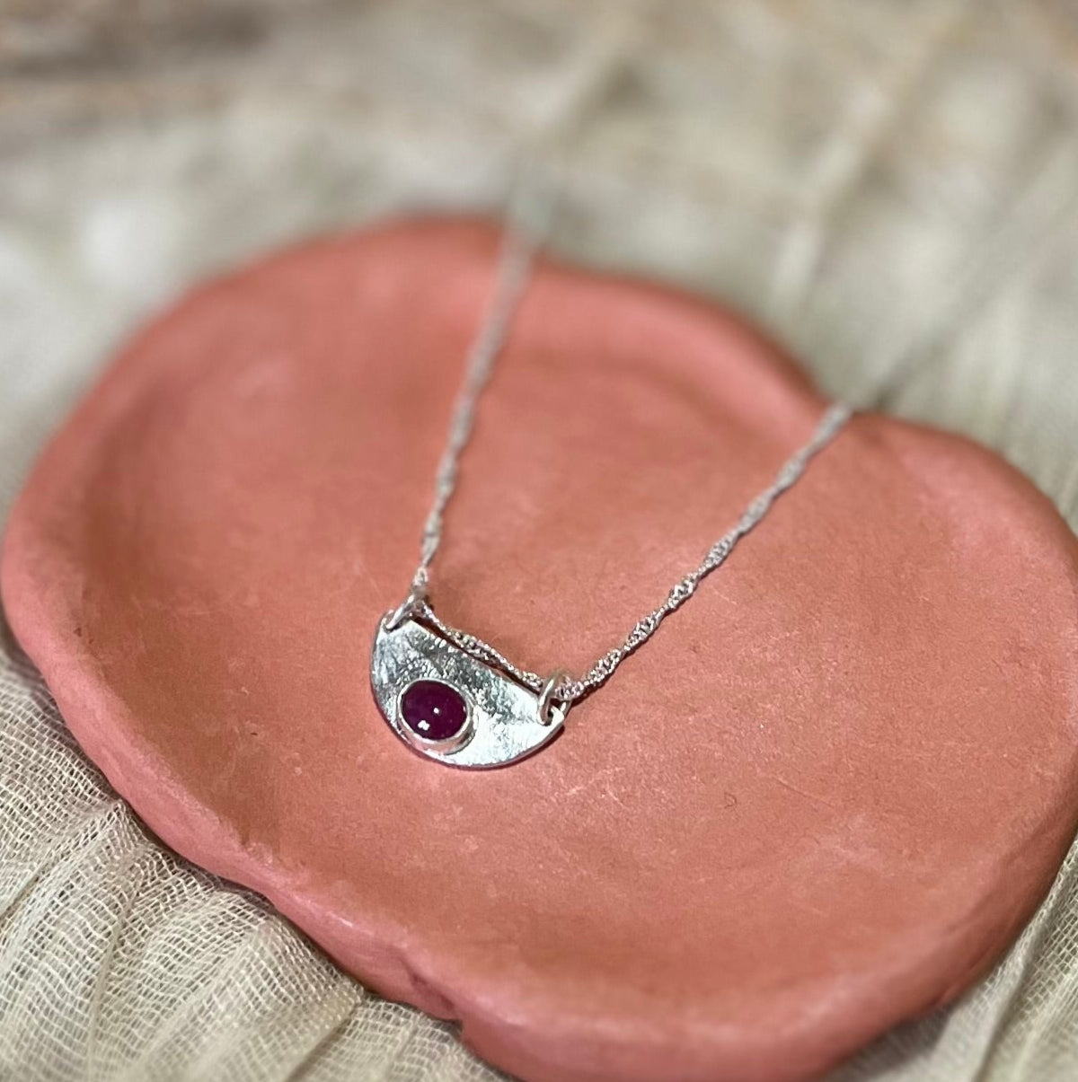 A sterling silver necklace with a textured pendant featuring an oval ruby stone, displayed on a terracotta dish.