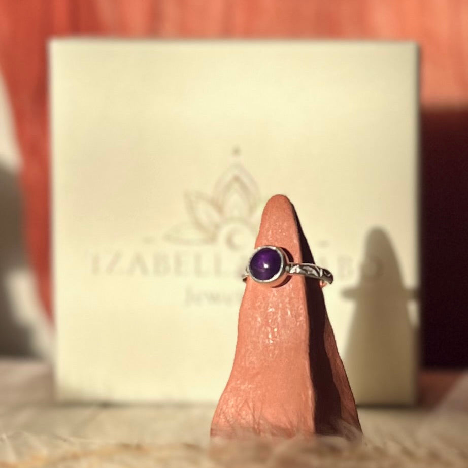 Silver ring with a purple gemstone on a finger, with a branded box in the background.