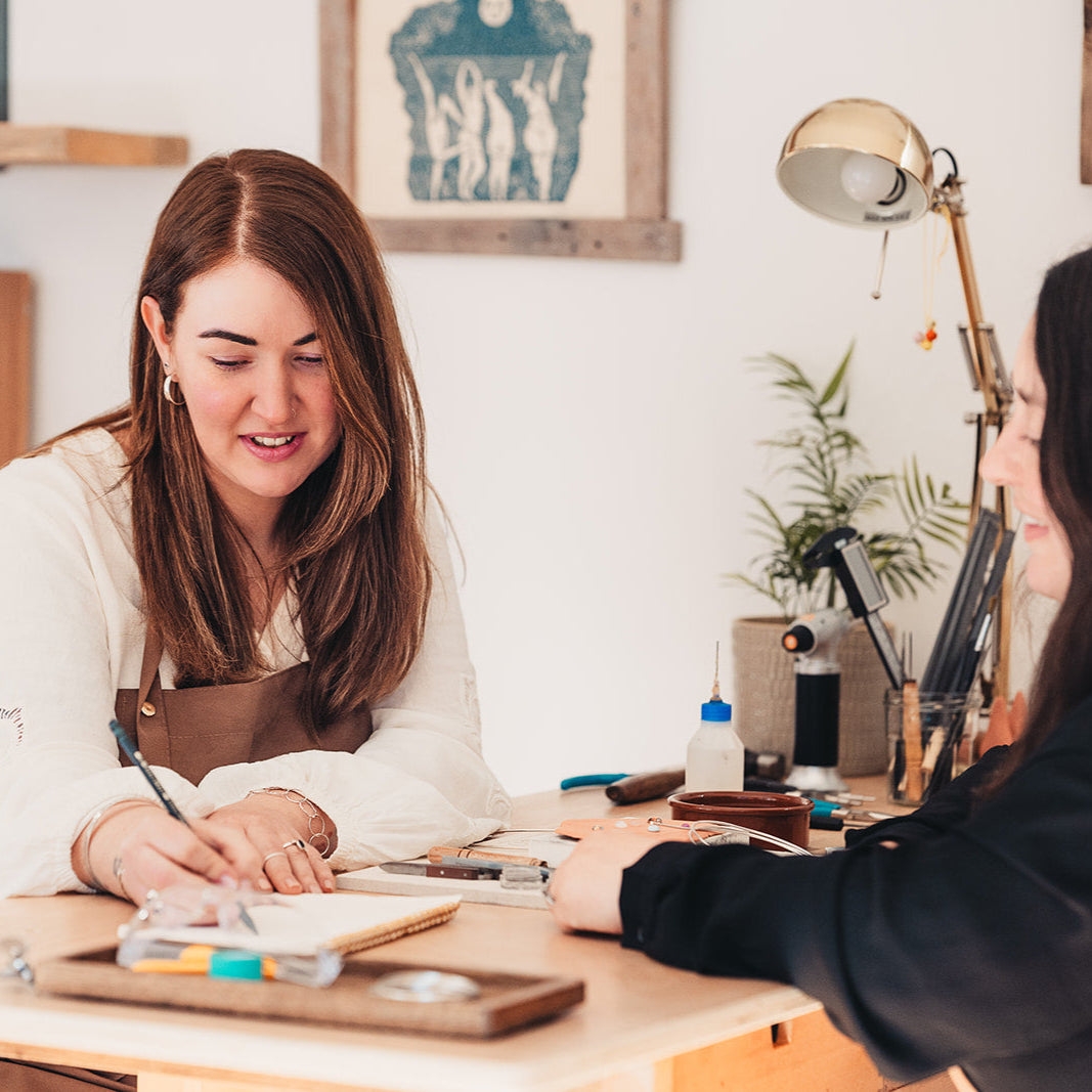 Two women in a workshop setting, one writing in a notebook while the other observes.