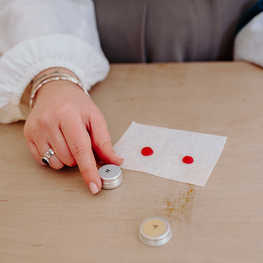 Person holding a small container next to two wax pieces on a piece of paper on a wooden table.