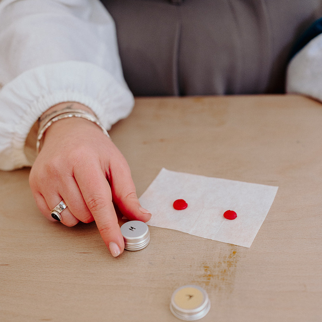 Person holding a small container next to two red dots on a piece of paper on a wooden table.