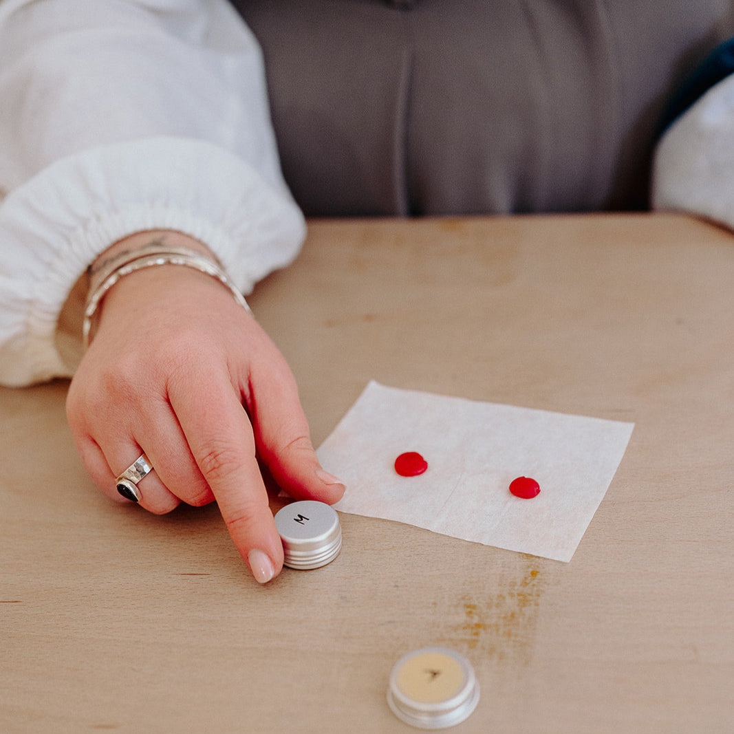 Person holding a small container next to two red dots on a piece of paper on a wooden table.