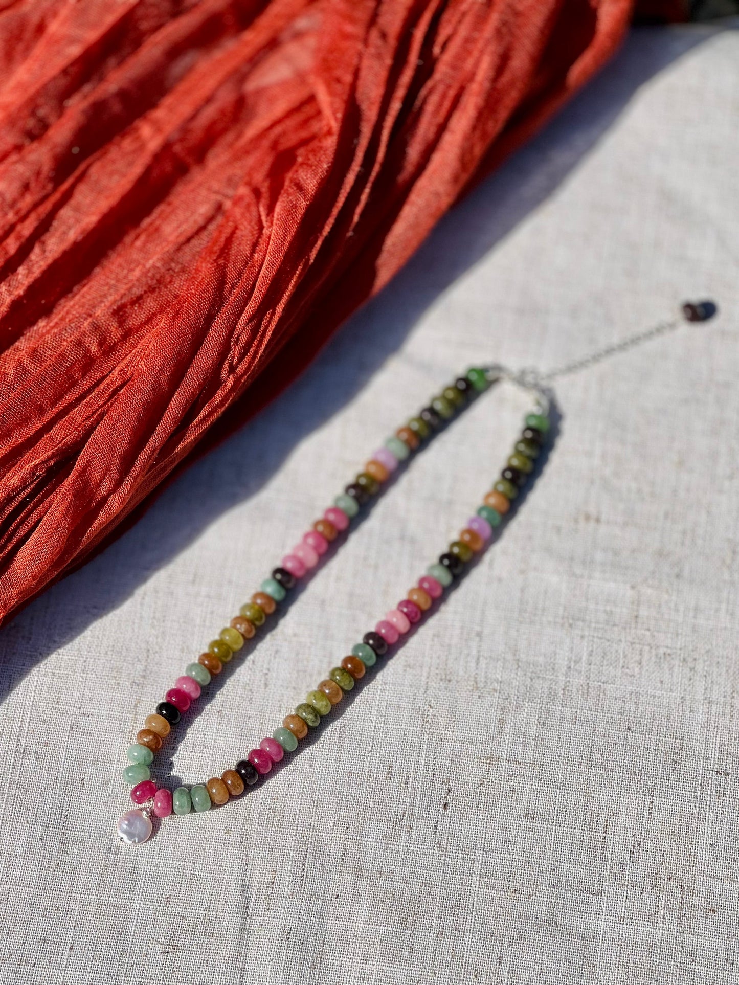 A beaded necklace with multicolored round and faceted beads, featuring a clear crystal pendant, laid out on a light beige linen surface next to a rust-orange textured fabric.