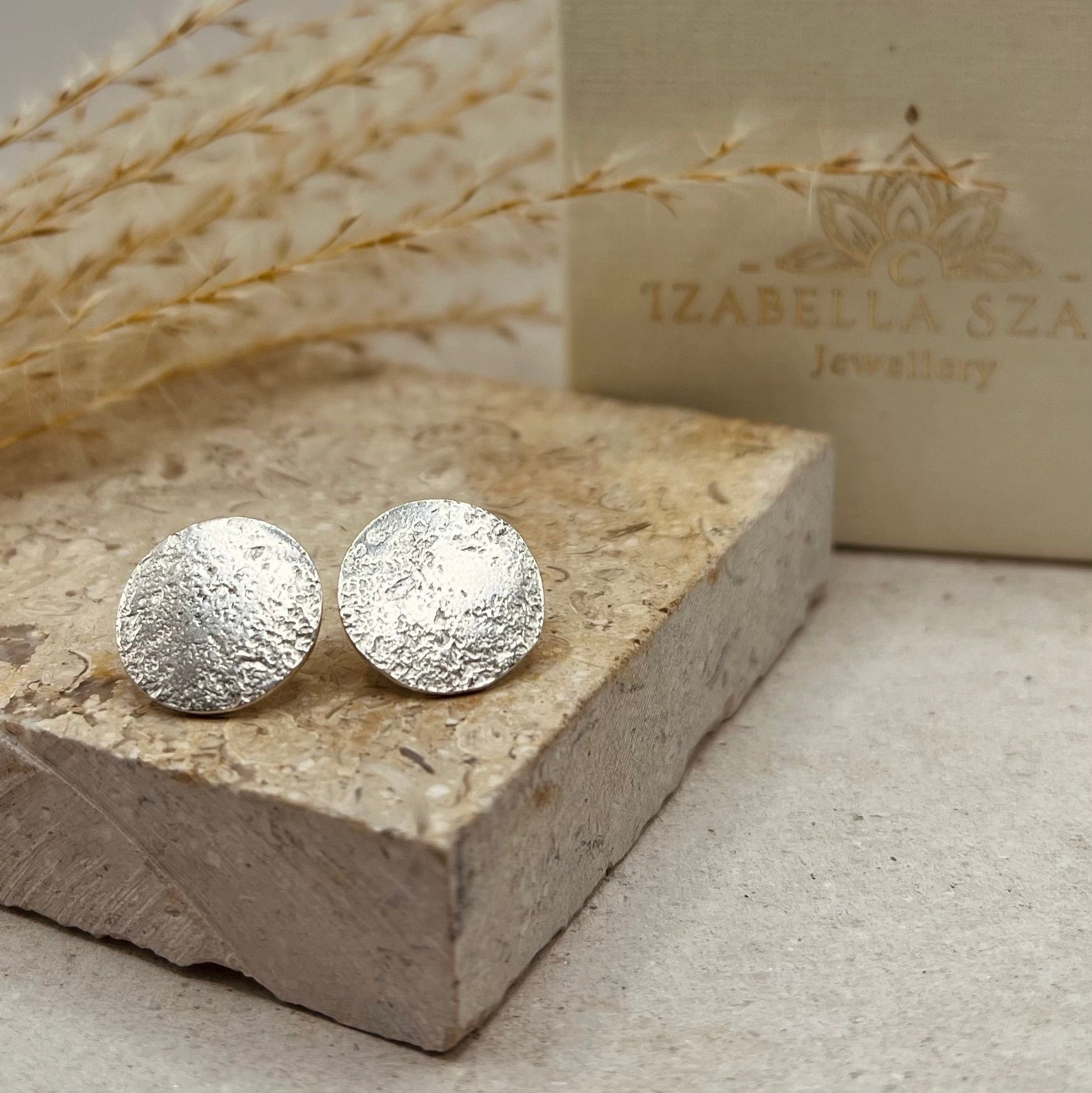 Silver round textured earrings on a stone surface with a box labeled 'Izabella Szabo Jewellery' in the background.