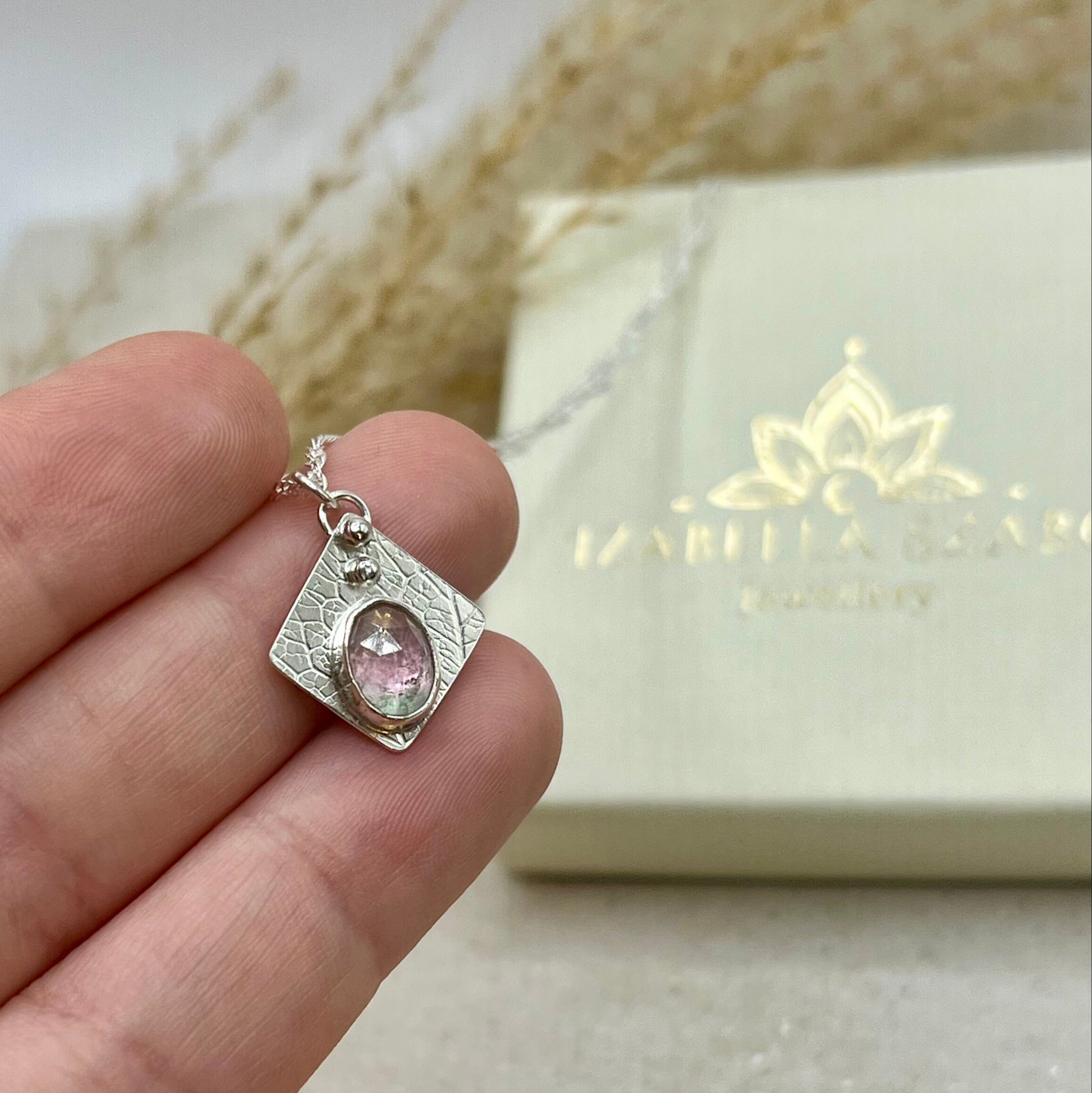 Silver pendant with pink gemstone held in a hand, brand Izabella Szabo Jewellery visible in the background.