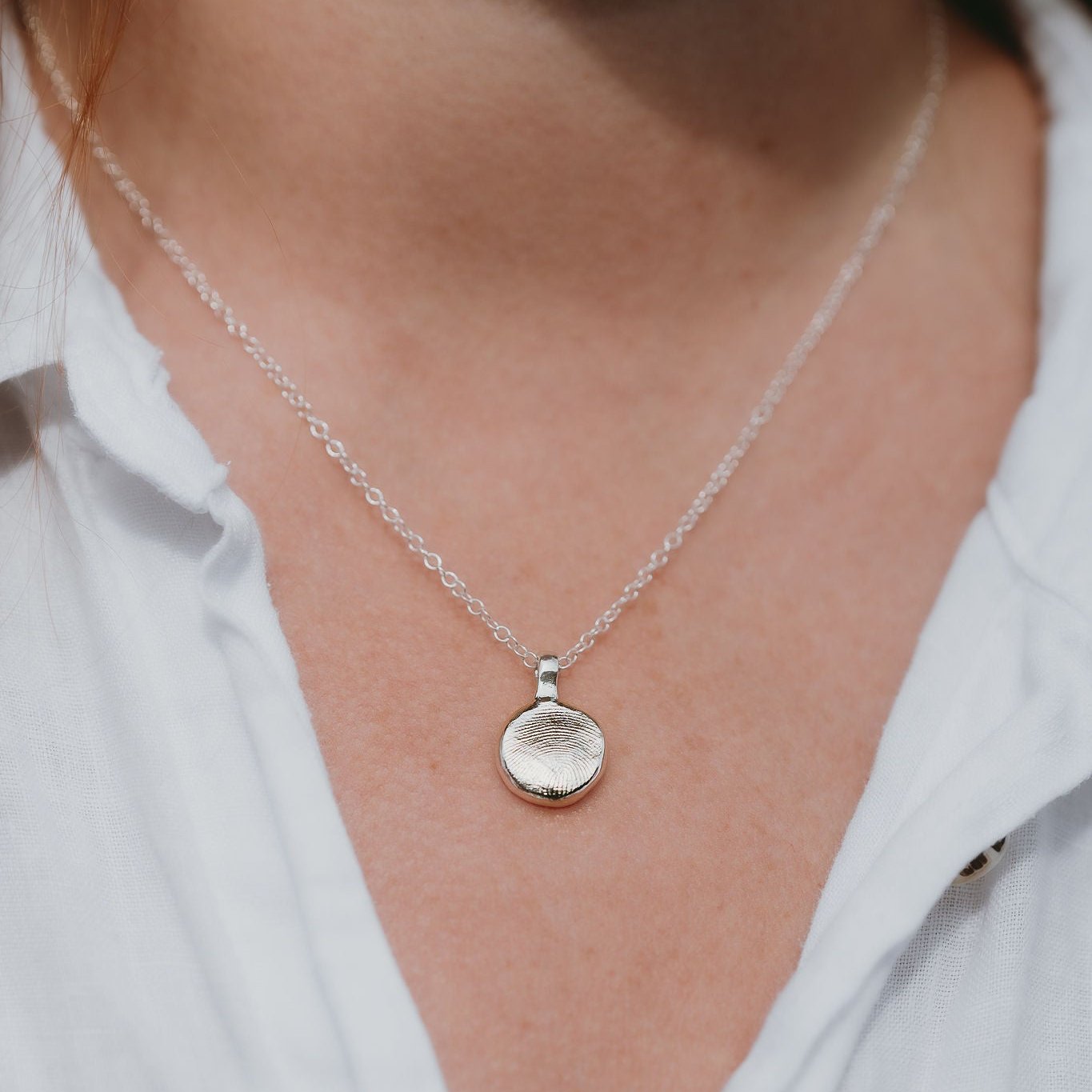 A close-up image of a silver fingerprint pendant necklace being worn, with a chain visible and a small, circular, silver pendant on the end bearing a fingerprint imprint.