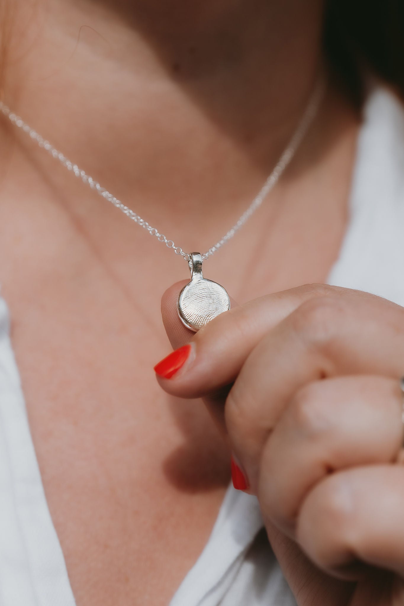 A close-up image of a silver fingerprint pendant necklace being worn, with a chain visible and a small, circular, silver pendant on the end bearing a fingerprint imprint.