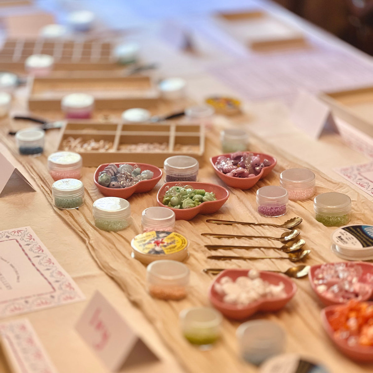 A styled tabletop display of small cosmetic jars, bowls of colorful powders or flakes, and gold-toned tools arranged on a wooden board in a softly blurred indoor setting.