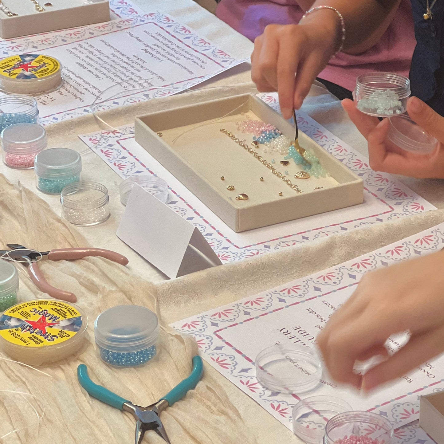 A close-up of hands crafting with small beads and tools on a table covered with papers, jars of colorful beads, pliers, and a tray containing a decorative design being assembled.