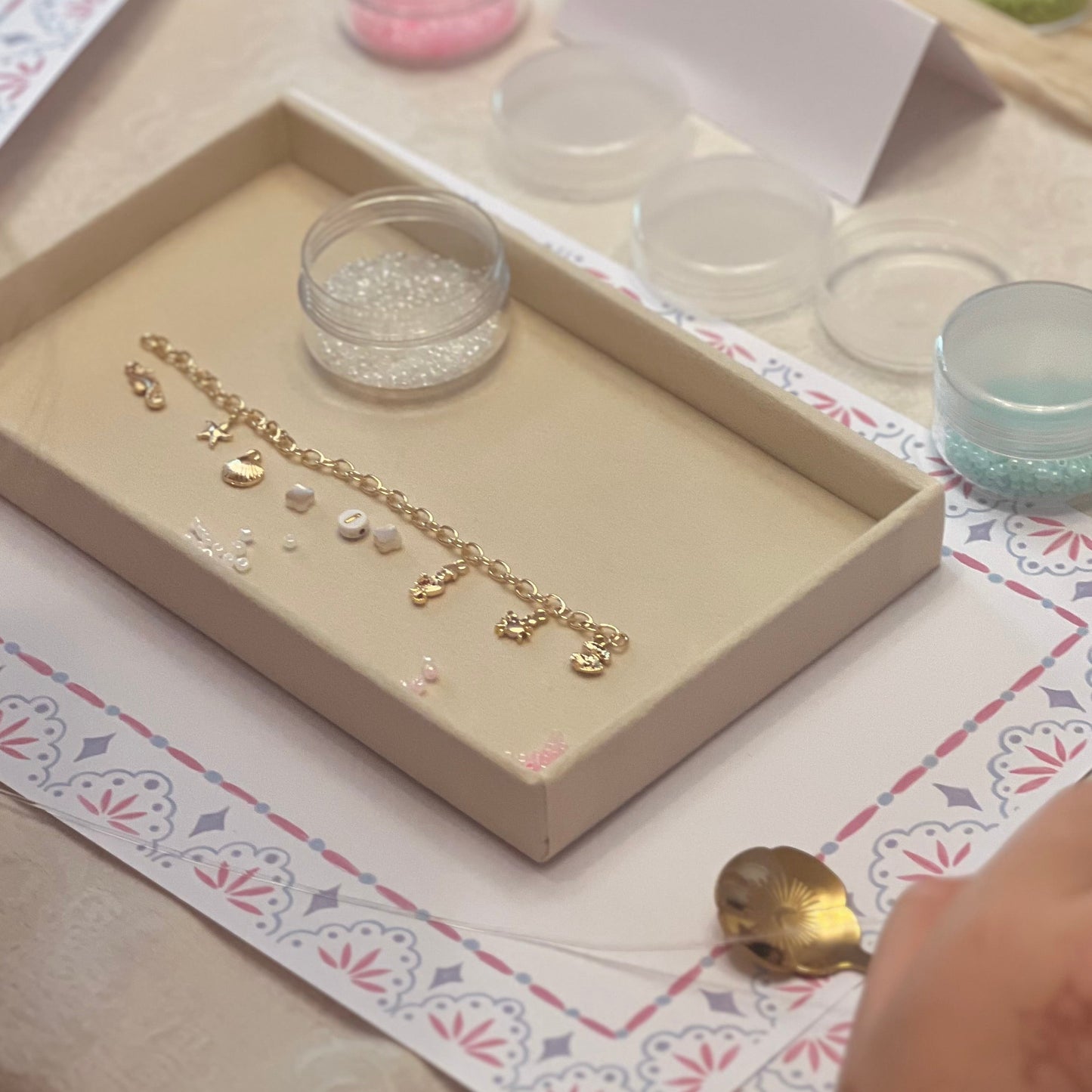 A gold charm bracelet with small animal and floral charms is displayed inside a beige jewelry tray on a decorated table. Small clear containers of beads or glitter, a white card, and a hand are visible around it.