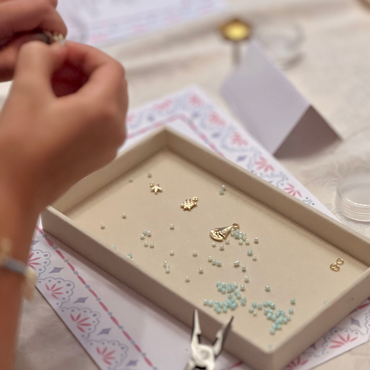 A close-up of hands crafting jewelry with small gold charms and light blue beads in a beige tray, with teal-handled pliers in the foreground on a patterned mat.