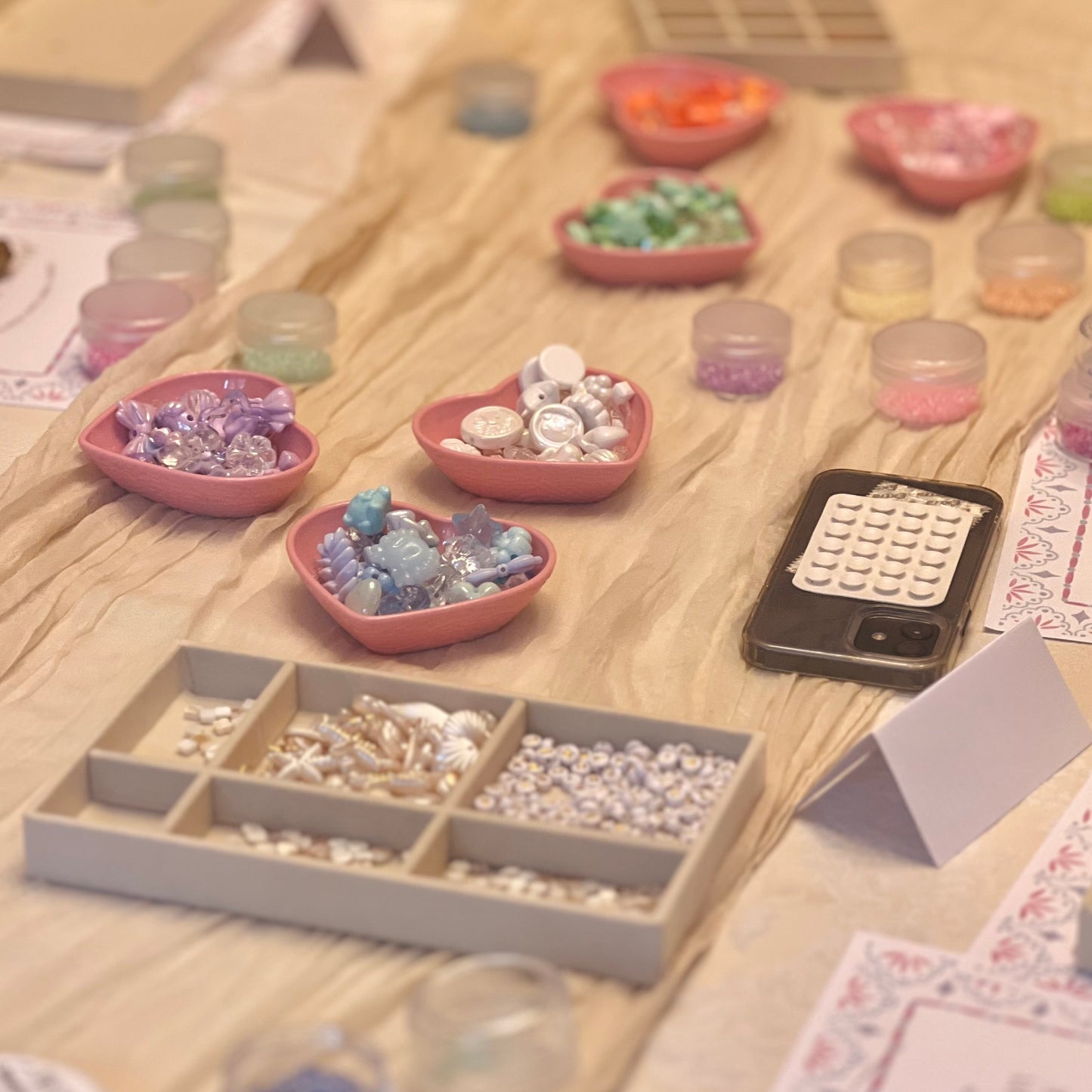 A styled tabletop display of craft supplies, including heart-shaped bowls filled with colorful beads, small clear jars, divided trays of rhinestones, and a black calculator on a light wood surface.