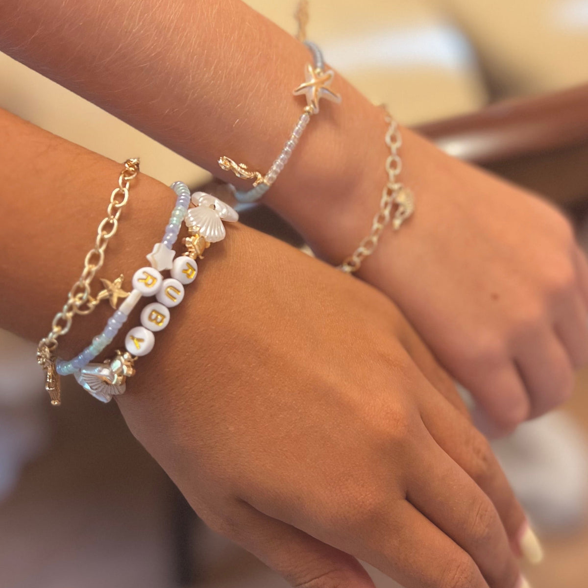 A close-up of a person's hands and wrists wearing multiple delicate bracelets, including gold-tone chain bracelets, a pearl bead bracelet, a light blue cord bracelet, and a starfish charm bracelet.