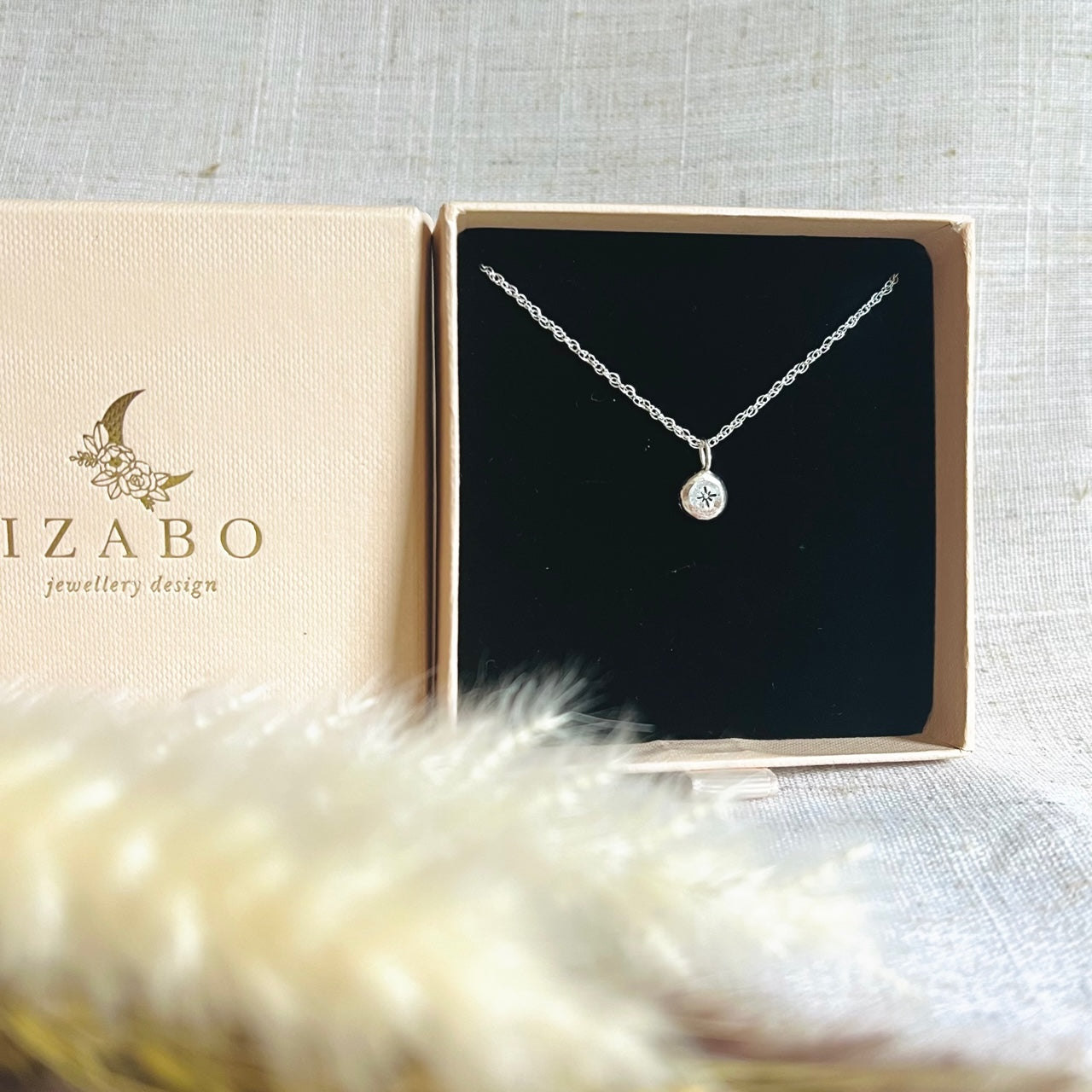 Little silver recycled nugget with star motif placed in it`s cream coloured IZABO branded gift box. The branded box has a gold IZABO jewellery design sign and a moon with flowers above. The top of the box and the bottom placed stood up next to each other on a cream coloured table cover, with dried foliage in front. 