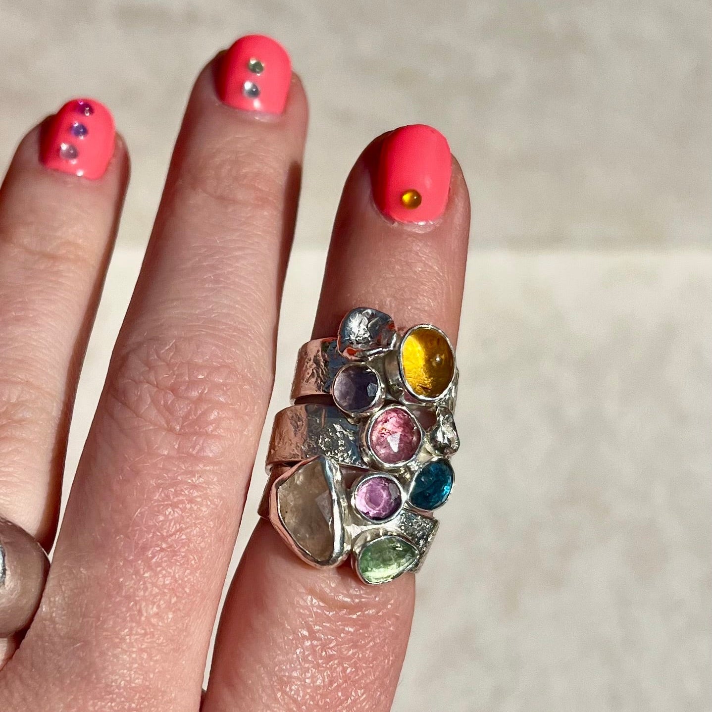 Hand wearing a ring with colorful gemstones on a neutral background