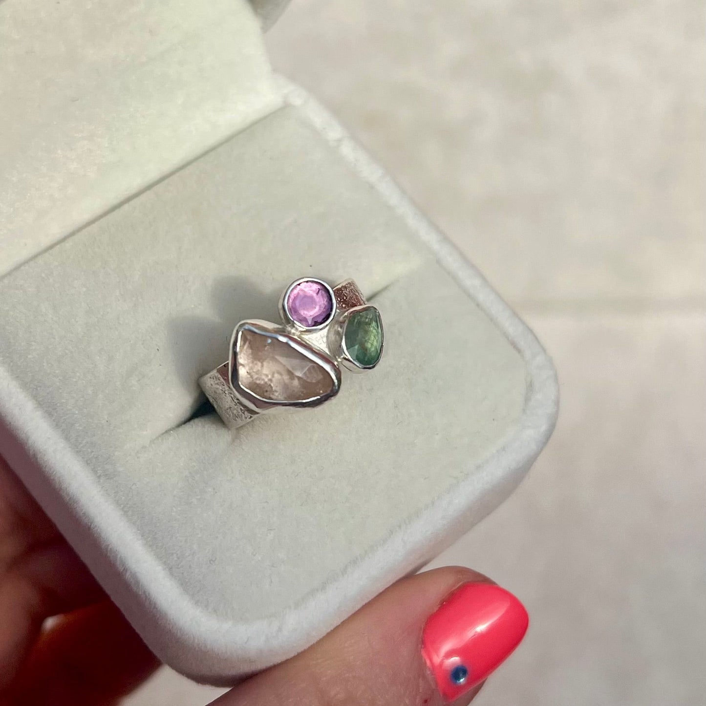 Silver ring with colorful stones in a white jewelry box held by a hand with pink nail polish.