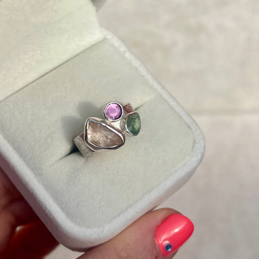 Silver ring with colorful stones in a white jewelry box held by a hand with pink nail polish.