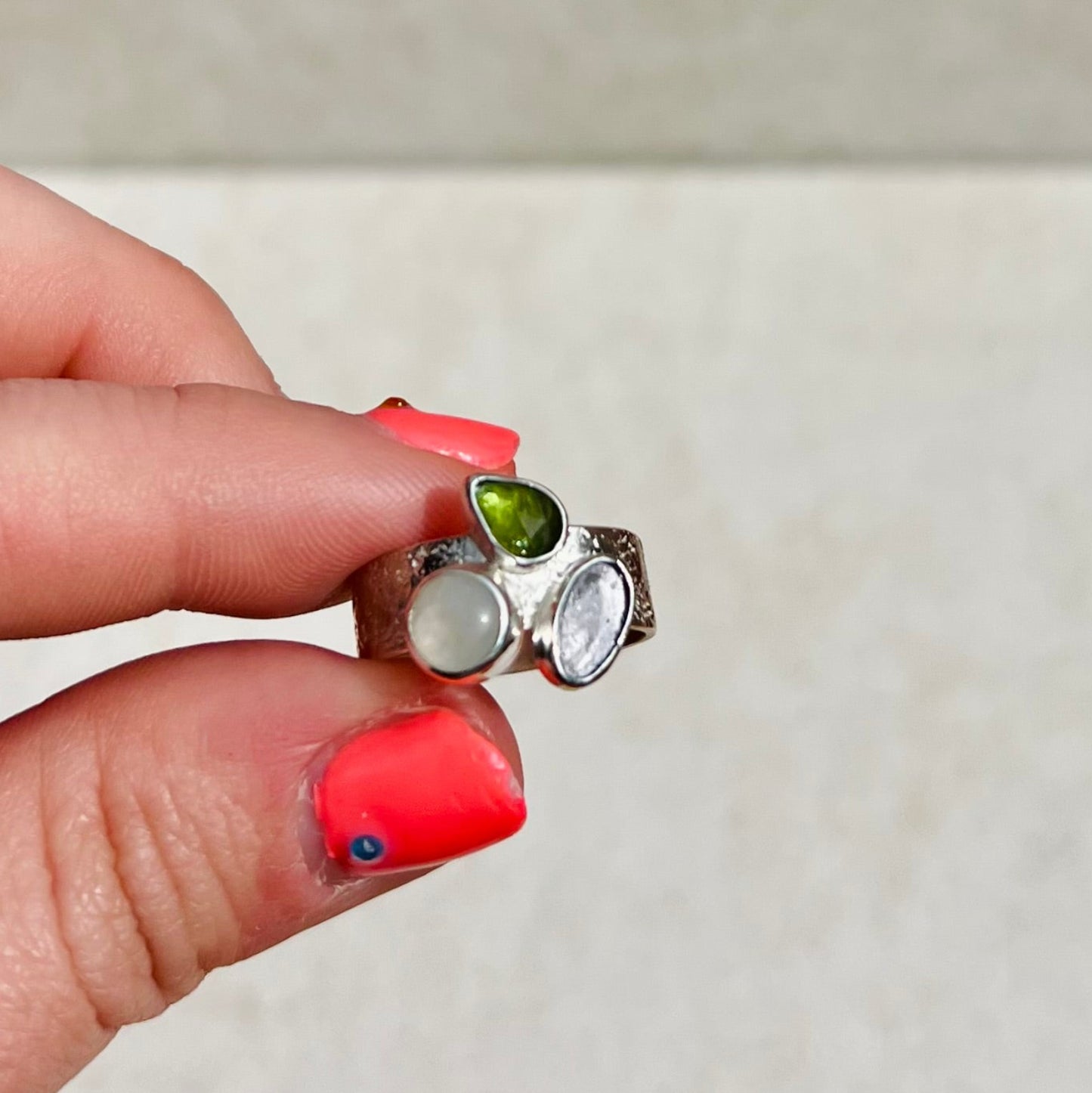 Close-up of a hand holding a silver ring with green and white gemstones against a neutral background.