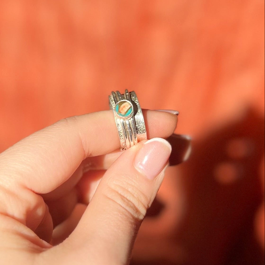 Silver ring with a colorful stone held between fingers against an orange background