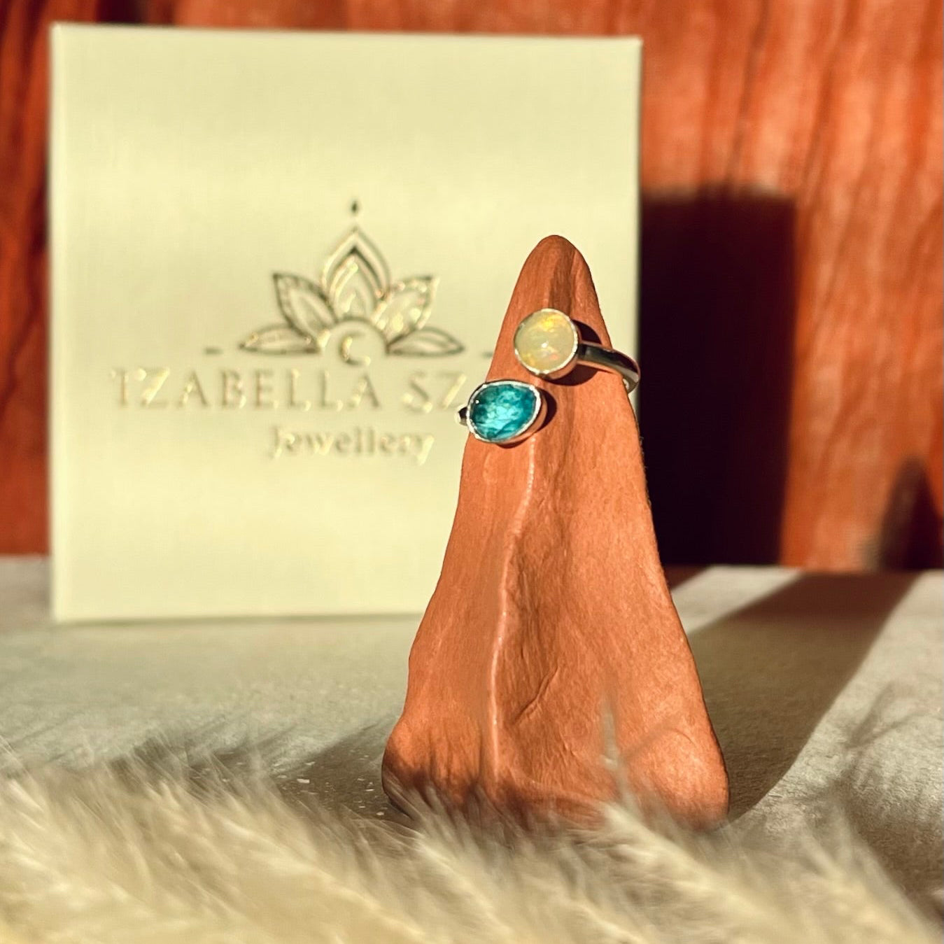 Silver ring with a blue gemstone on a wooden block, with Izabella Szabo Jewellery box in the background.