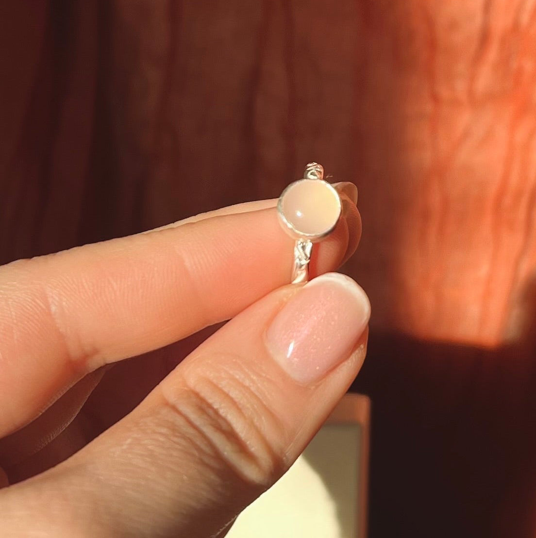 Close-up of a hand holding a silver ring with a peach-colored gemstone against a blurred background.
