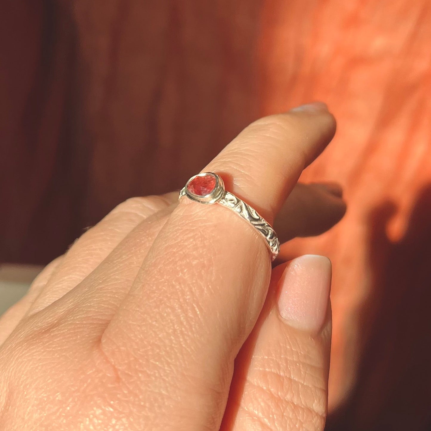 Close-up of a hand wearing a silver ring with a red stone against a blurred background