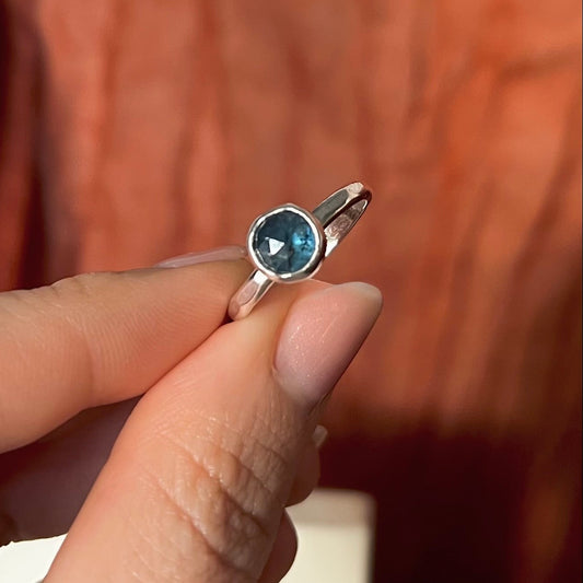Silver ring with a blue gemstone held between fingers against a blurred background
