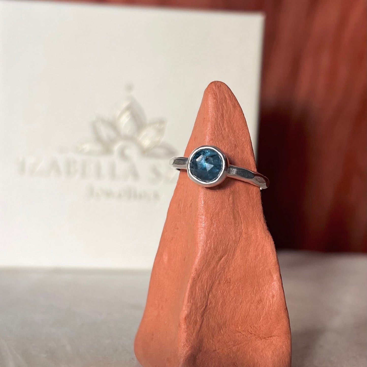 Silver ring with a blue gemstone on a wooden stand, with a blurred background