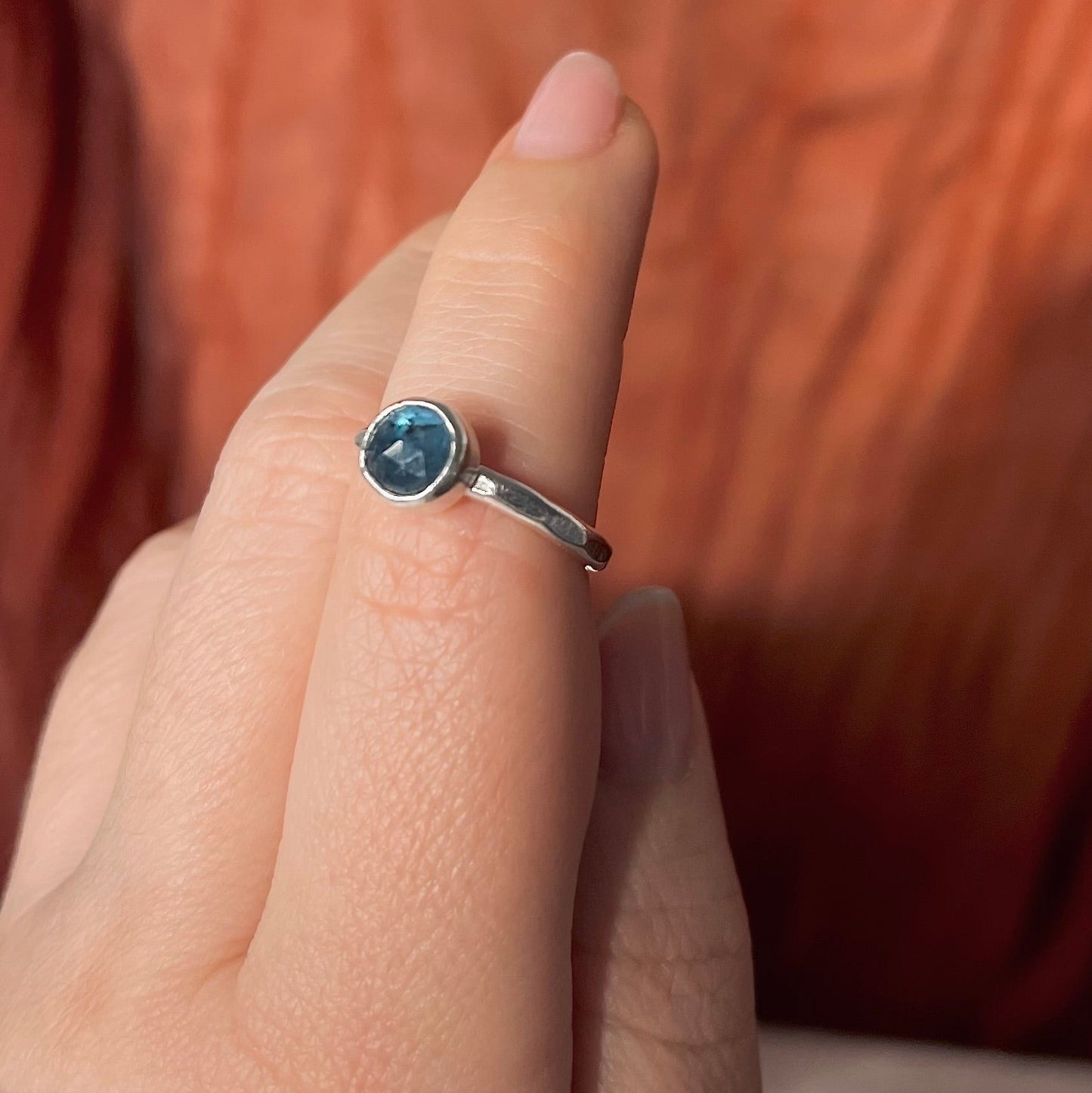 Hand wearing a silver ring with a blue gemstone against a warm-toned fabric background