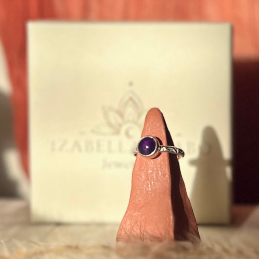 Silver ring with a purple gemstone on a finger, with a branded box in the background.