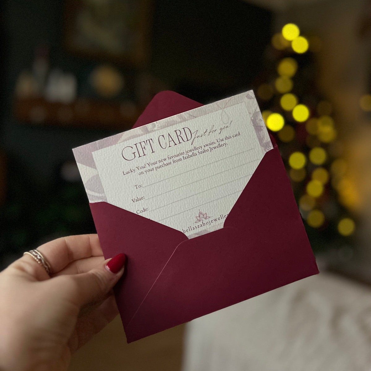A hand with red nail polish holds a white gift card with elegant script text, tucked inside a deep burgundy envelope. The background is a softly blurred indoor setting with warm yellow bokeh lights, suggesting a cozy holiday or home environment.