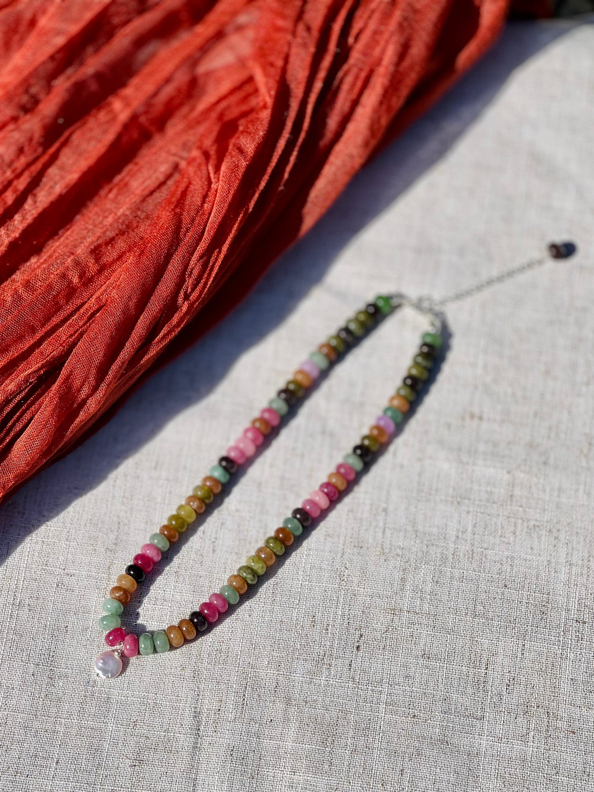 A beaded necklace with multicolored round and faceted beads, featuring a clear crystal pendant, laid out on a light beige linen surface next to a rust-orange textured fabric.