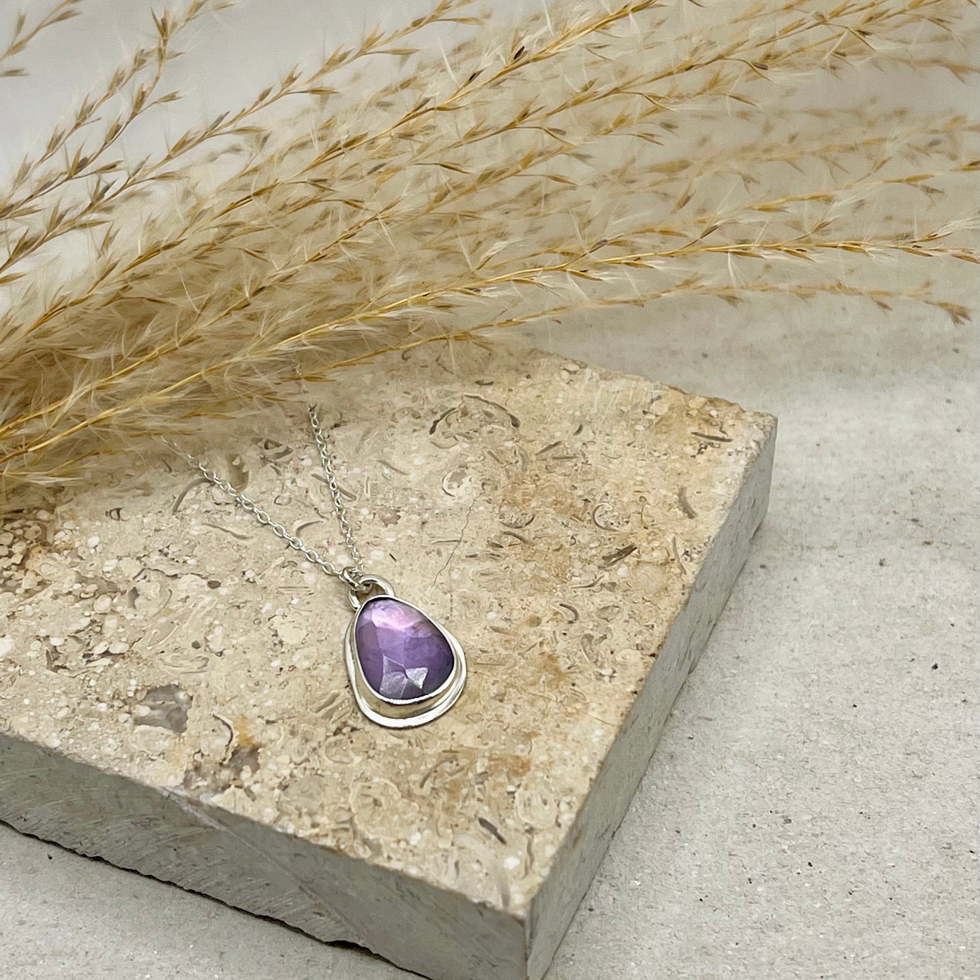 Purple gemstone pendant on a silver chain, placed on a concrete surface with dried grasses.